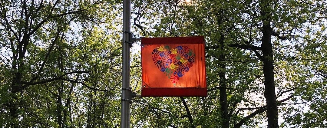 Photo of outside light with Indigenous flag commemorating the 215 children, it is an orange flag with hand prints forming a heart. behind is lush green trees.