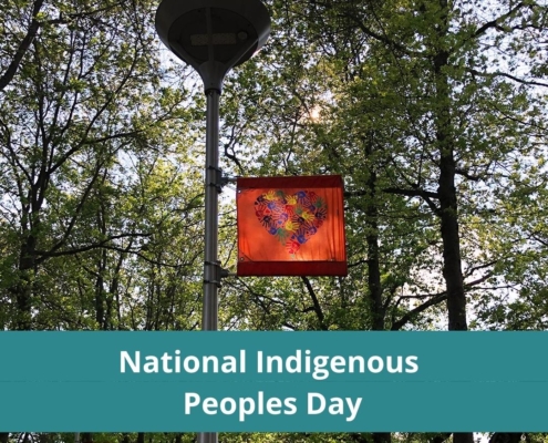 Photo of outside light with Indigenous flag commemorating the 215 children, it is an orange flag with hand prints forming a heart. behind is lush green trees.